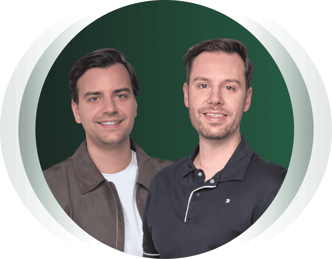 Zwei Männer stehen nebeneinander vor einem grünen Hintergrund und lächeln beide in die Kamera. Einer trägt eine braune Jacke und ein weißes Hemd, der andere ein schwarzes Hemd mit Kragen.