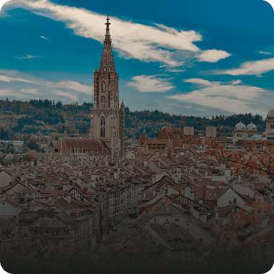 Ein hoher Kirchturm erhebt sich über einer dichten Ansammlung von Ziegeldächern mit Bäumen und einem teilweise bewölkten Himmel im Hintergrund.