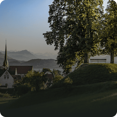 Auf einem grasbewachsenen Hügel steht eine Bank, umgeben von Bäumen, mit Blick auf ein Dorf mit einem Kirchturm und Bergen im Hintergrund.