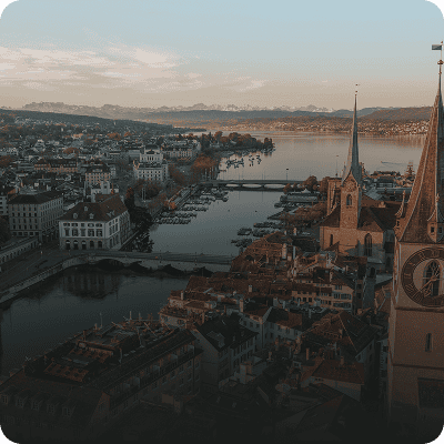 Luftaufnahme einer europäischen Stadt mit einem Fluss, historischen Gebäuden, Kirchtürmen und Bergen im Hintergrund unter klarem Himmel.