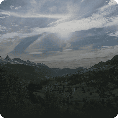 Berglandschaft mit schneebedeckten Gipfeln in der Ferne, ein Tal mit verstreuten Bäumen und Häusern und dramatischen Wolken, die die Sonne teilweise verdecken.