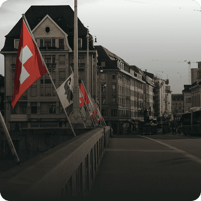 Eine Reihe Schweizer Flaggen säumt unter bewölktem Himmel eine Brücke vor historischen Gebäuden in einer Stadtstraße.