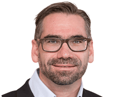 Ein Mann mit kurzen braunen Haaren, Bart und Brille trägt ein Hemd mit Kragen und eine dunkle Jacke und blickt direkt in die Kamera.