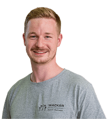 Ein Mann mit kurzem hellbraunem Haar und gestutztem Bart, der ein graues T-Shirt mit der Marke Wacker trägt und in die Kamera lächelt.