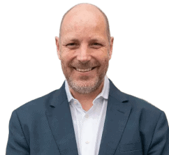 Ein glatzköpfiger Mann mit Bart, dunklem Jackett und weißem Hemd lächelt vor einem schlichten Hintergrund in die Kamera.