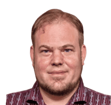 Ein Mann mit kurzem hellbraunem Haar, gestutztem Bart und gemustertem Hemd blickt vor einem schlichten Hintergrund direkt in die Kamera.