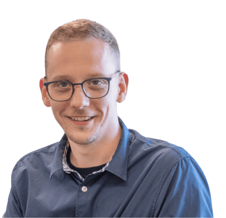 Ein Mann mit kurzen Haaren und Brille, der ein blaues Hemd mit Knöpfen trägt, lächelt, vor einem neutralen Hintergrund.