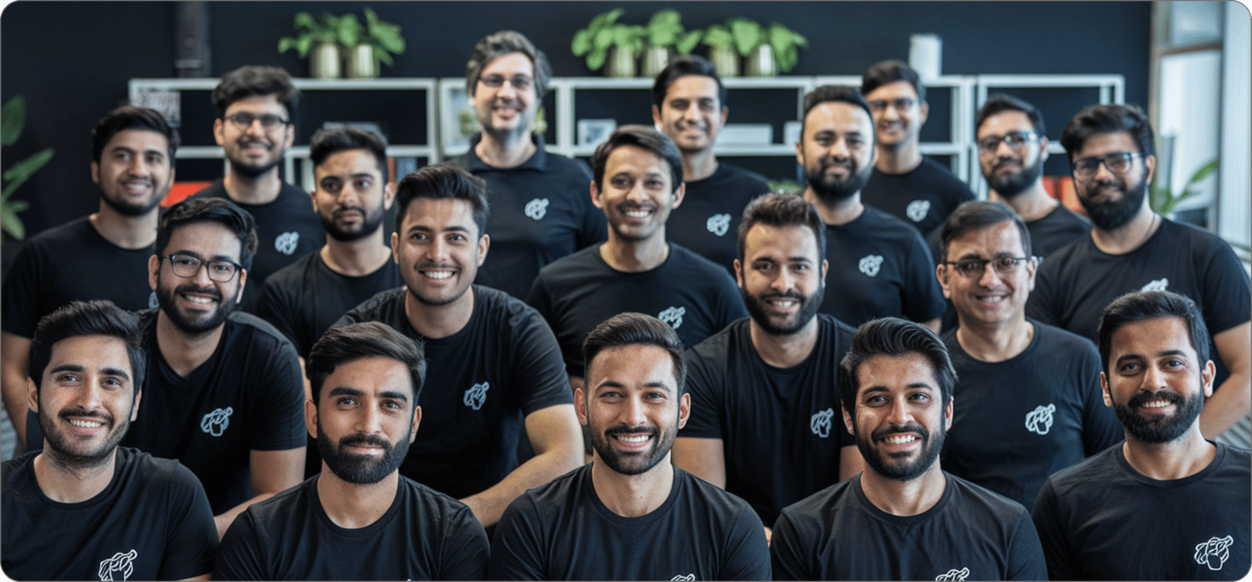 Eine Gruppe von neunzehn Personen in passenden schwarzen T-Shirts posiert für ein Foto im Innenbereich, im Hintergrund sind Regale und Topfpflanzen zu sehen.