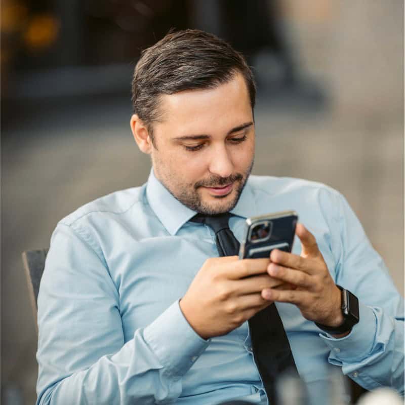 Ein Mann mit hellblauem Hemd und schwarzer Krawatte sitzt da und schaut auf sein Smartphone. Er wirkt konzentriert und beschäftigt, möglicherweise mit der Durchsicht von Unterlagen für einen bevorstehenden SEO-Workshop.