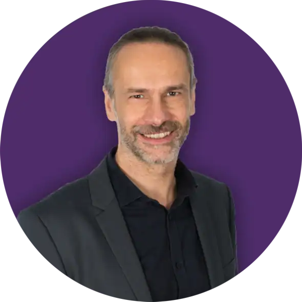Ein Mann mit kurzen grauen Haaren und Bart, der eine dunkle Anzugsjacke und ein schwarzes Hemd trägt, lächelt selbstbewusst vor einem lila Hintergrund und ist bereit, einen SEO-Workshop zu leiten.