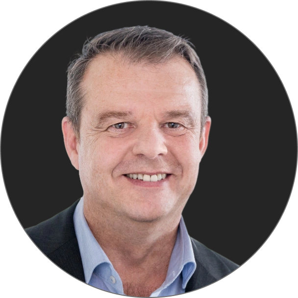 Mann mittleren Alters mit kurzen braunen Haaren, der ein hellblaues Hemd mit Kragen und einen dunklen Blazer trägt, lächelnd, vor einem schlichten dunklen Hintergrund.