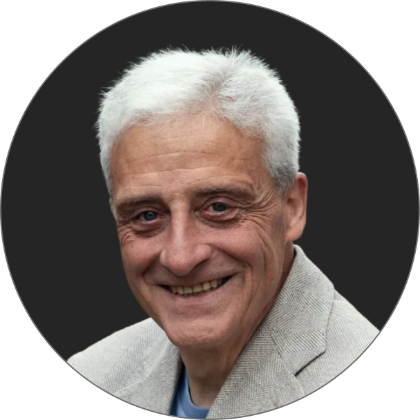 Ein älterer Mann mit kurzen weißen Haaren und einer hellgrauen Jacke lächelt in die Kamera vor einem dunklen runden Hintergrund.