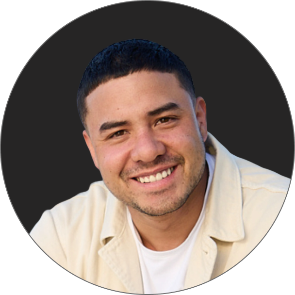 Ein Mann mit kurzen dunklen Haaren und Gesichtsbehaarung lächelt in die Kamera. Er trägt eine helle Jacke über einem weißen Hemd und steht vor einem dunklen, runden Hintergrund.